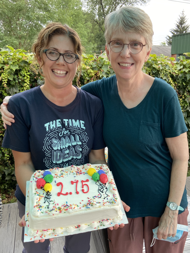 Photo of two women standing side by side embracing and smiling. The woman on the left is holding a sheet cake with 