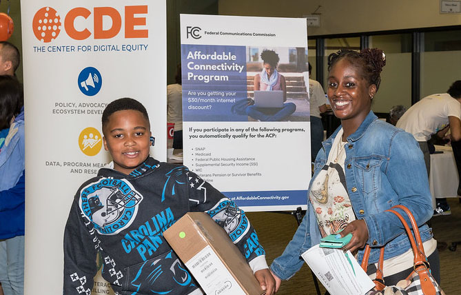A Black boy, about 10 years old, stands in front of a sign holding a computer box smiling. To his right, a Black woman holds his hand and smiles.