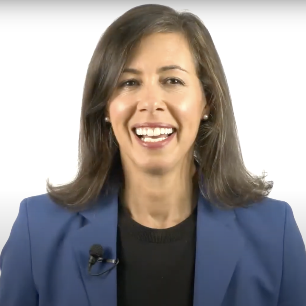 A woman with brown hair, blue suit, and black shirt smiles