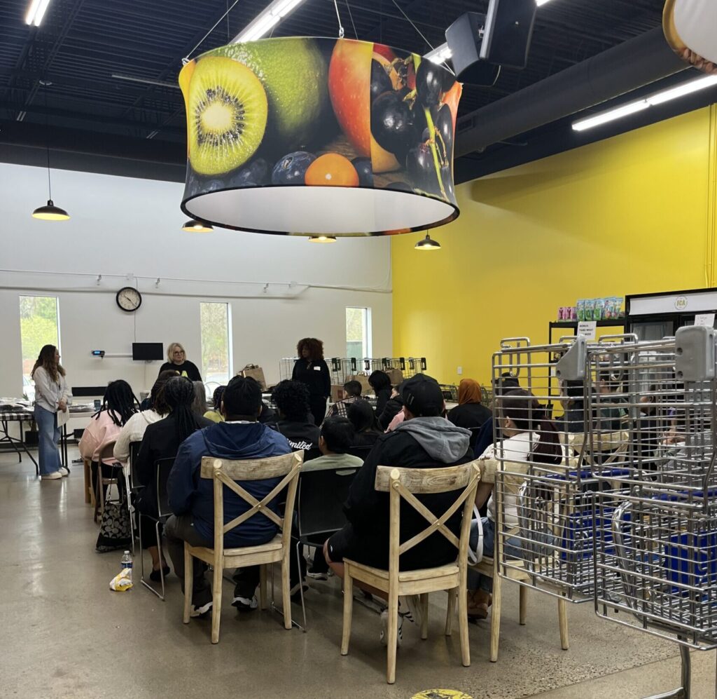 Approximately 20 community members seated in chairs, facing 2 speakers at the front of the room. Images of fruit on the central overhead light fixture.